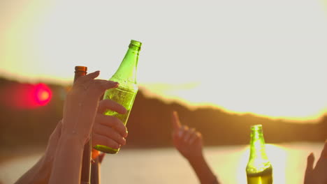 Young-students-are-dancing-with-hands-up-and-clink-beer-from-colorful-bottles.-This-is-crazy-party-at-sunset-on-the-river-coast.